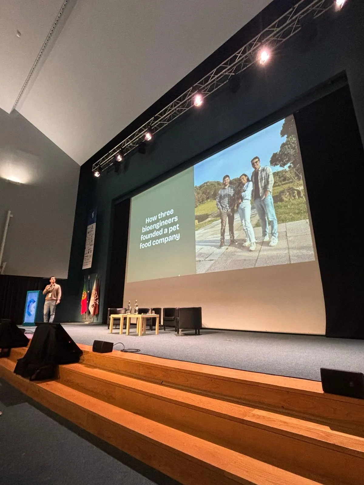 A large auditorium stage with a slide reading “How three bioengineers founded a pet food company”, and João speaking at the lectern.