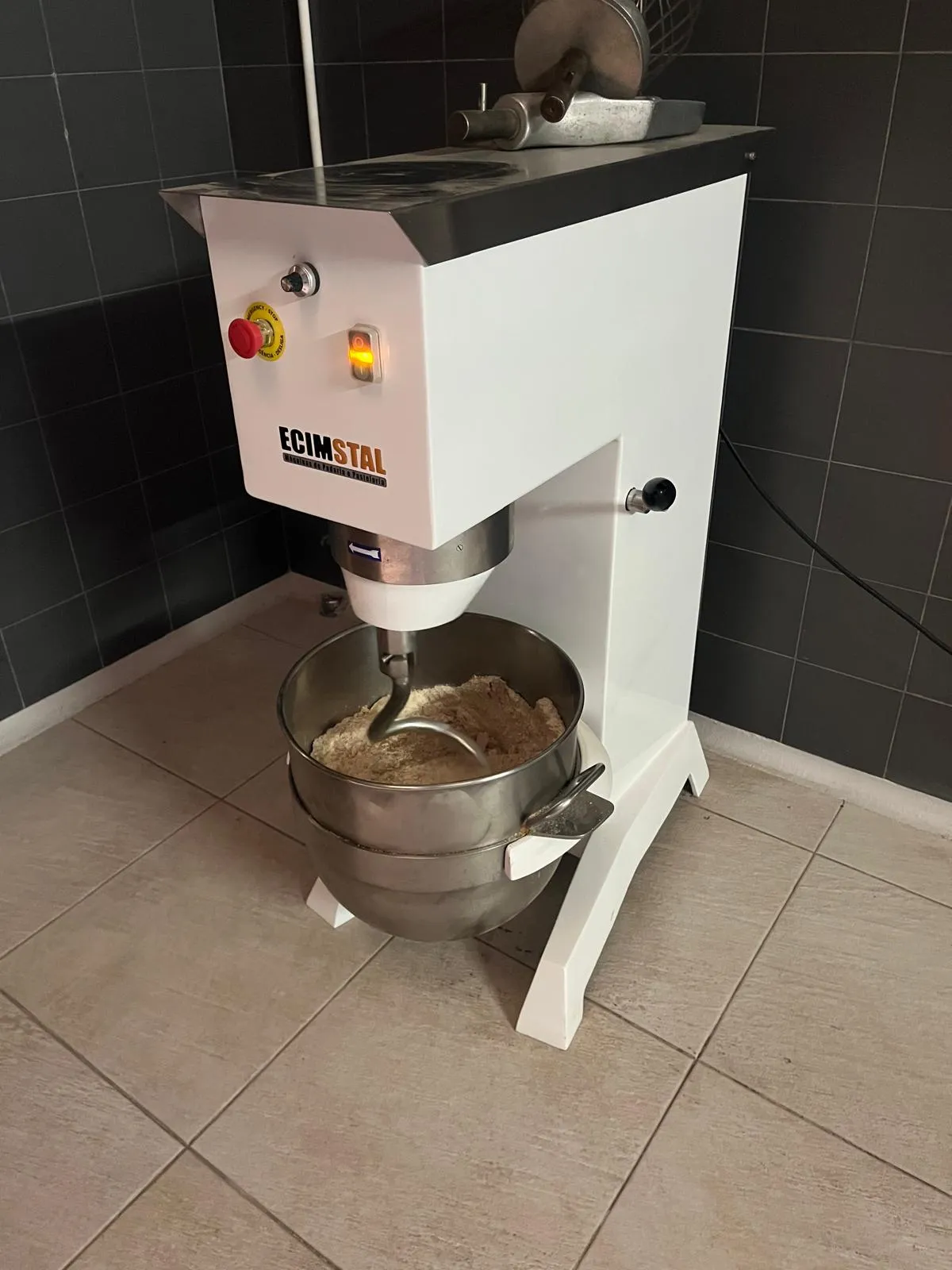 An industrial mixer with a stainless steel bowl of mixed ingredients, standing in a tiled production room.