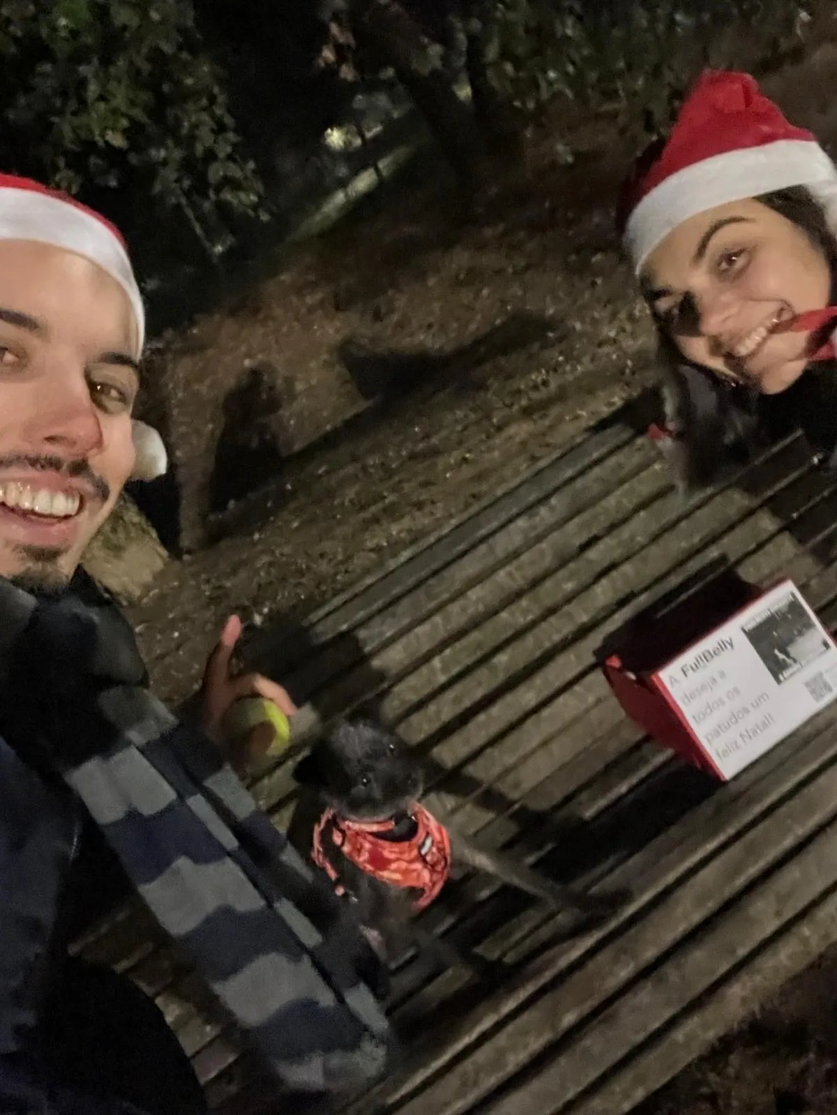 A night-time photo of two people in Santa hats on a park bench with a dog and a red Full Belly gift box.