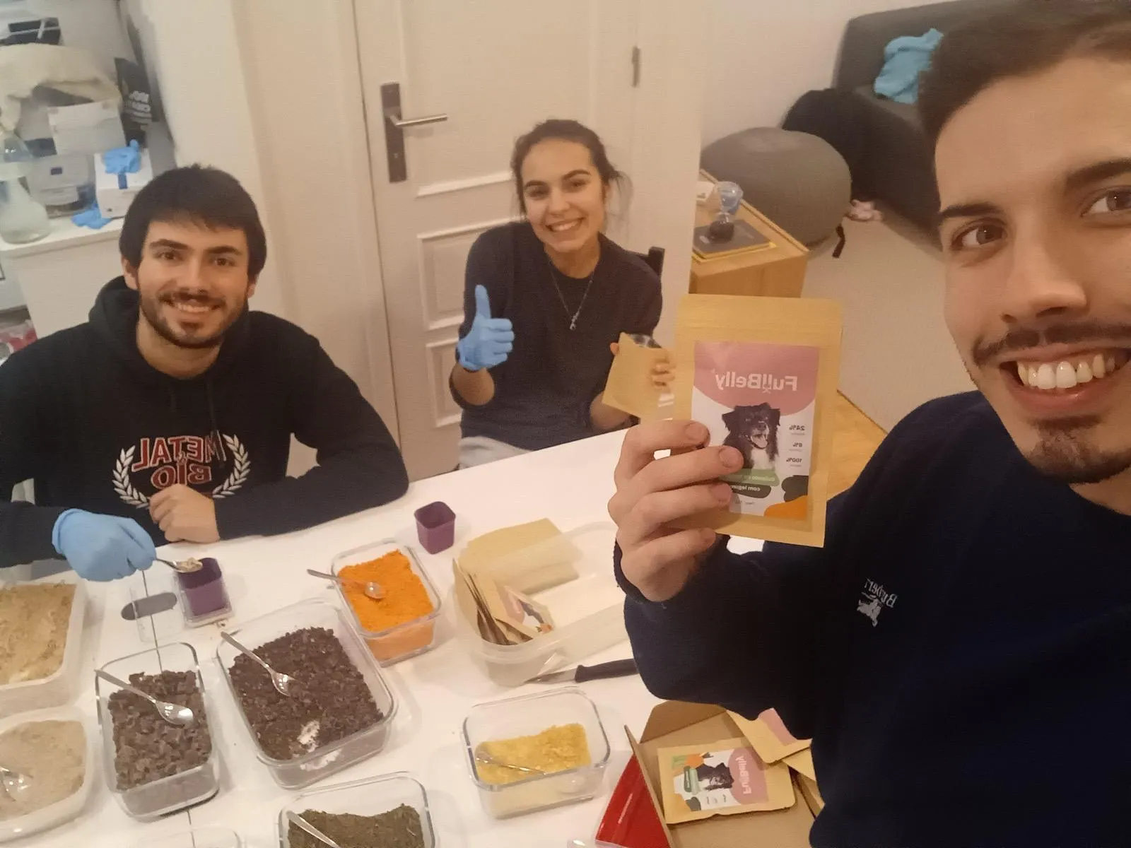 Three people around a table covered with trays of dehydrated ingredients, holding up an early Full Belly pouch.