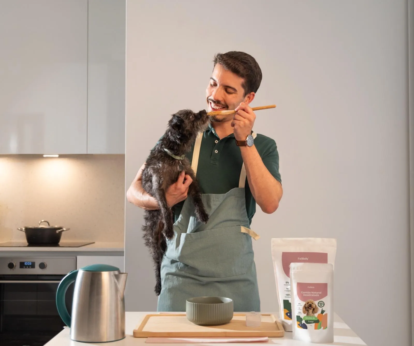 João holding Suki in a kitchen, offering her a wooden spoon, with two Full Belly product pouches on the counter.