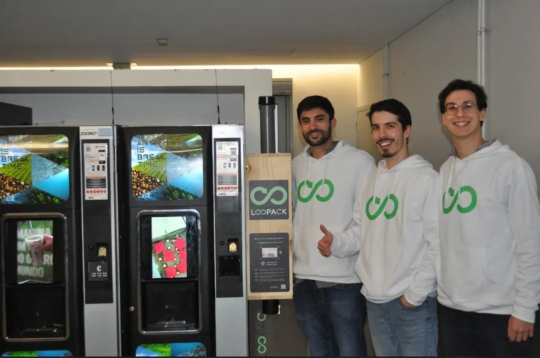 Three people in Loopack hoodies standing beside the reusable-cup dispenser, next to the coffee vending machines it was installed against.